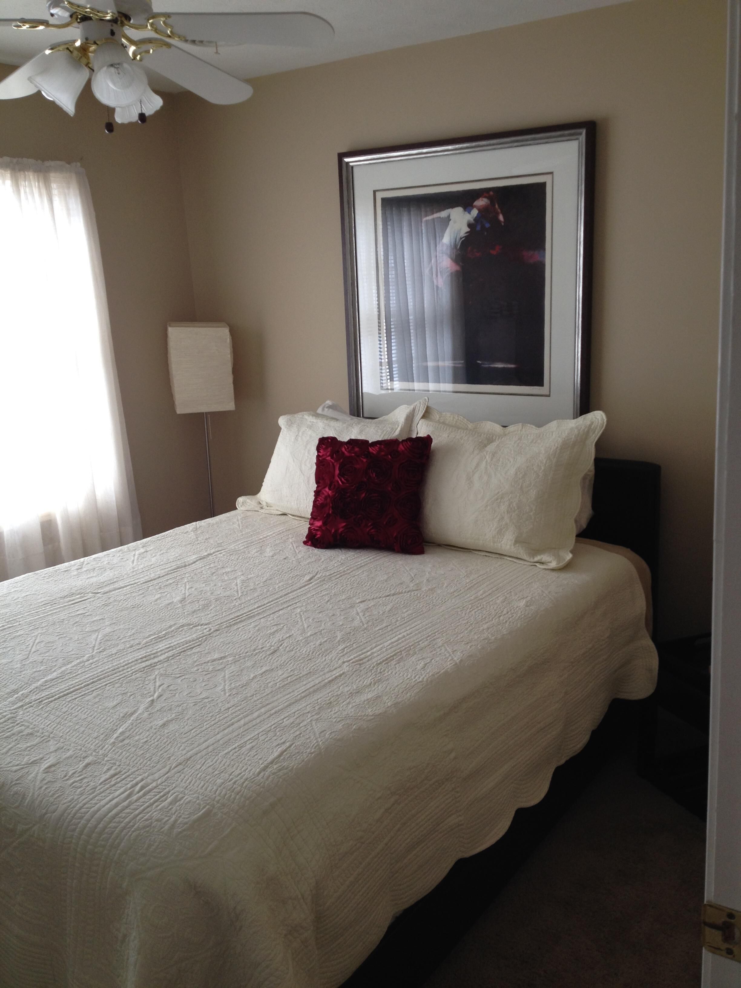 Una cama bien tendida con sábanas blancas y un cojín rojo decorativo, un cuadro en la pared y un ventilador de techo.