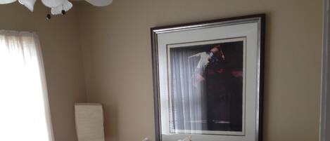 Una cama bien tendida con sábanas blancas y un cojín rojo decorativo, un cuadro en la pared y un ventilador de techo.