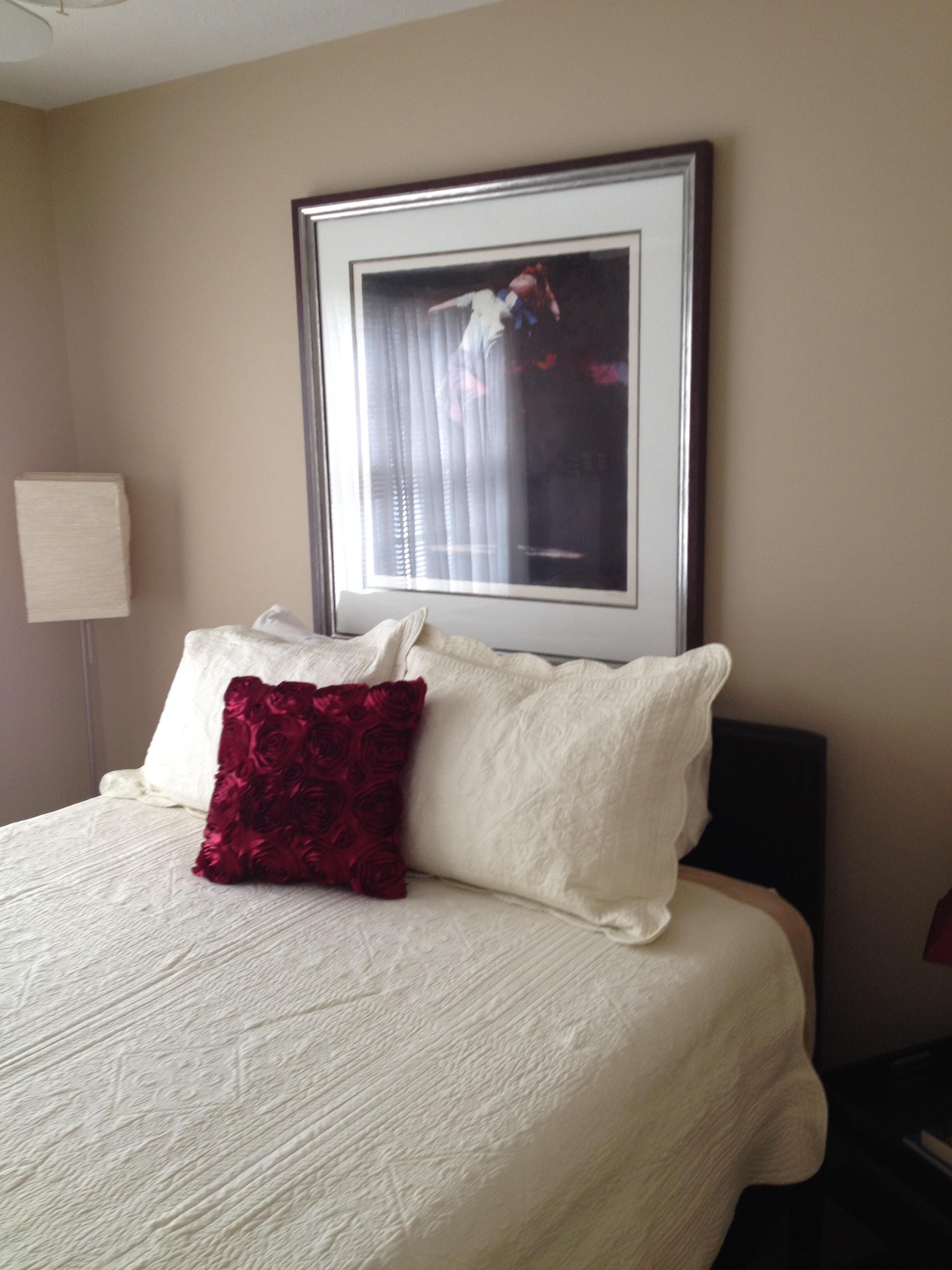 A neatly made bed with white and red pillows, a framed picture on the wall, and a bedside lamp.