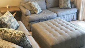Ein Wohnzimmer mit einem grauen Ecksofa, mehreren Kissen mit Muster, einem grauen Hocker, einem Landschaftsbild und einer Stehlampe.