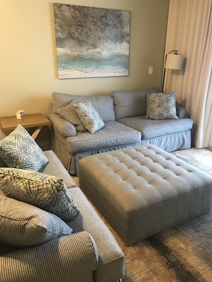 A living room with a grey sectional sofa, multiple patterned throw pillows, a grey ottoman, a painting of a landscape, and a floor lamp.