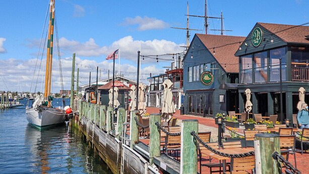 Ein Restaurant am Wasserufer mit Außenbereich, ein Segelboot liegt in der Nähe, und ein klarer, blauer Himmel.