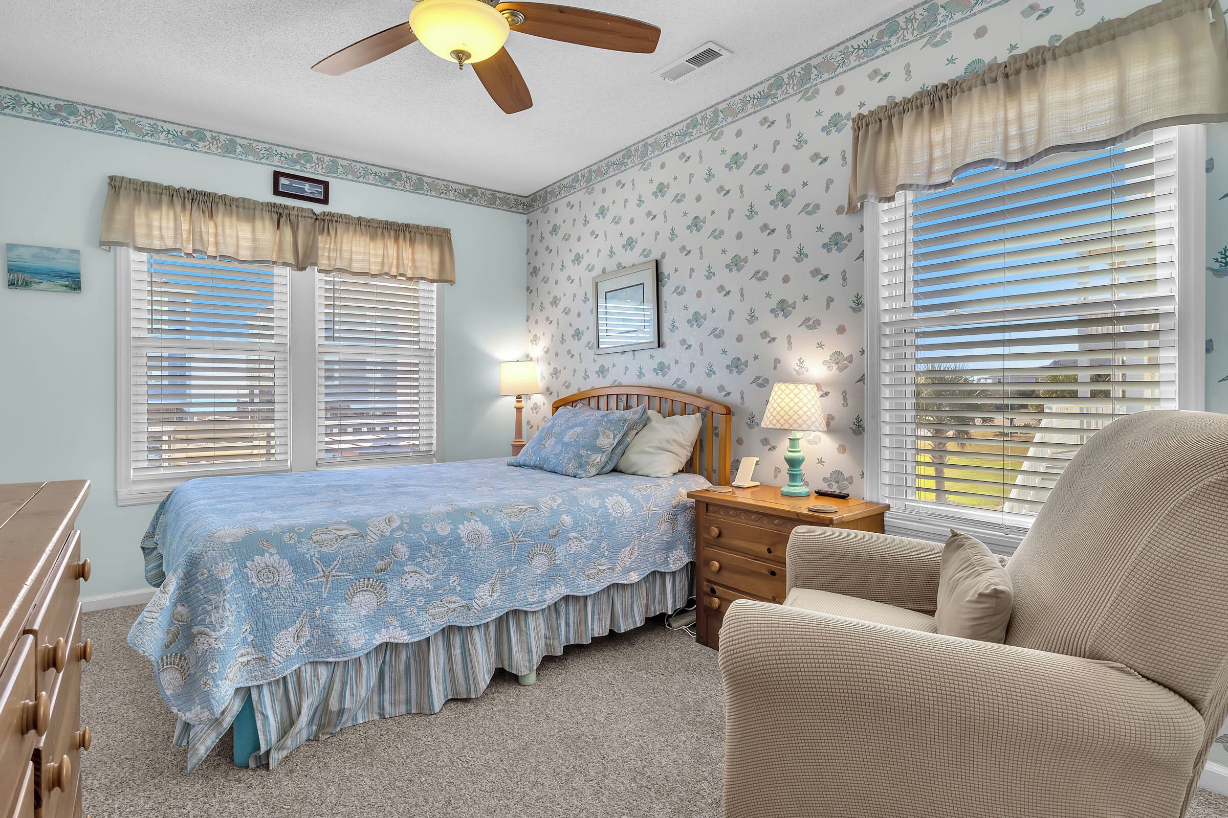 A bedroom with a bed, two windows with blinds, a ceiling fan, a nightstand, a chair, and a dresser.