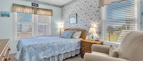 A bedroom with a bed, two windows with blinds, a ceiling fan, a nightstand, a chair, and a dresser.