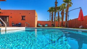 Une piscine d’un bleu éclatant, avec des chaises longues et un parasol rouge, entourée de palmiers et d’un bâtiment rouge.