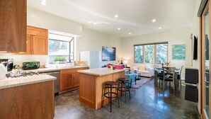 A modern kitchen with wooden cabinets, a central island with stools, a dining area with a table and chairs, and a living room with a sofa and a TV.