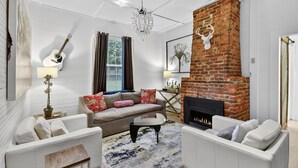 A living room with a brick fireplace, white sofas, a chandelier, and a guitar on the wall. - Blue Ridge- Hot Tub - EV Outlet -Outdoor Oasis (Blue Ridge)