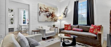 Ett modernt vardagsrum med en beige soffa, en brun soffa med röda kuddar, en vit fåtölj och ett soffbord i glas. På väggen hänger en stor, abstrakt tavla, och i taket finns en kristallkrona. En gitarr står lutad mot väggen.