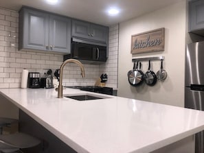 A modern kitchen with grey cabinets, a stainless steel refrigerator, a white countertop, and a wall-mounted pot rack.