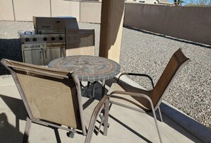 An outdoor patio with a round table and two wicker chairs, a grill, and a stone wall in the background. - snowbirds email of call for winter pricing<br>951-741-7590 occapt@hotmail.com (Lake Havasu City)