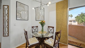 A dining area with a glass table, four wooden chairs, a vase with flowers, and a modern chandelier. - Dessert Oasis - 2BR Desert Breeze Retreat with Pool - jacuzzi- Gym (Phoenix)