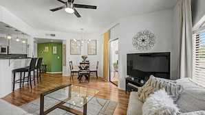 A modern living room with a glass coffee table, a sofa, a TV, and a dining area with chairs and a table. - Dessert Oasis - 2BR Desert Breeze Retreat with Pool - jacuzzi- Gym (Phoenix)
