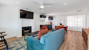 A modern living room with a blue sofa, a flat-screen TV mounted on the wall, a fireplace, and a kitchen area in the background. - Oceanviews, Steps to the Sand, Short Walk to the Village! A/C (Carlsbad)