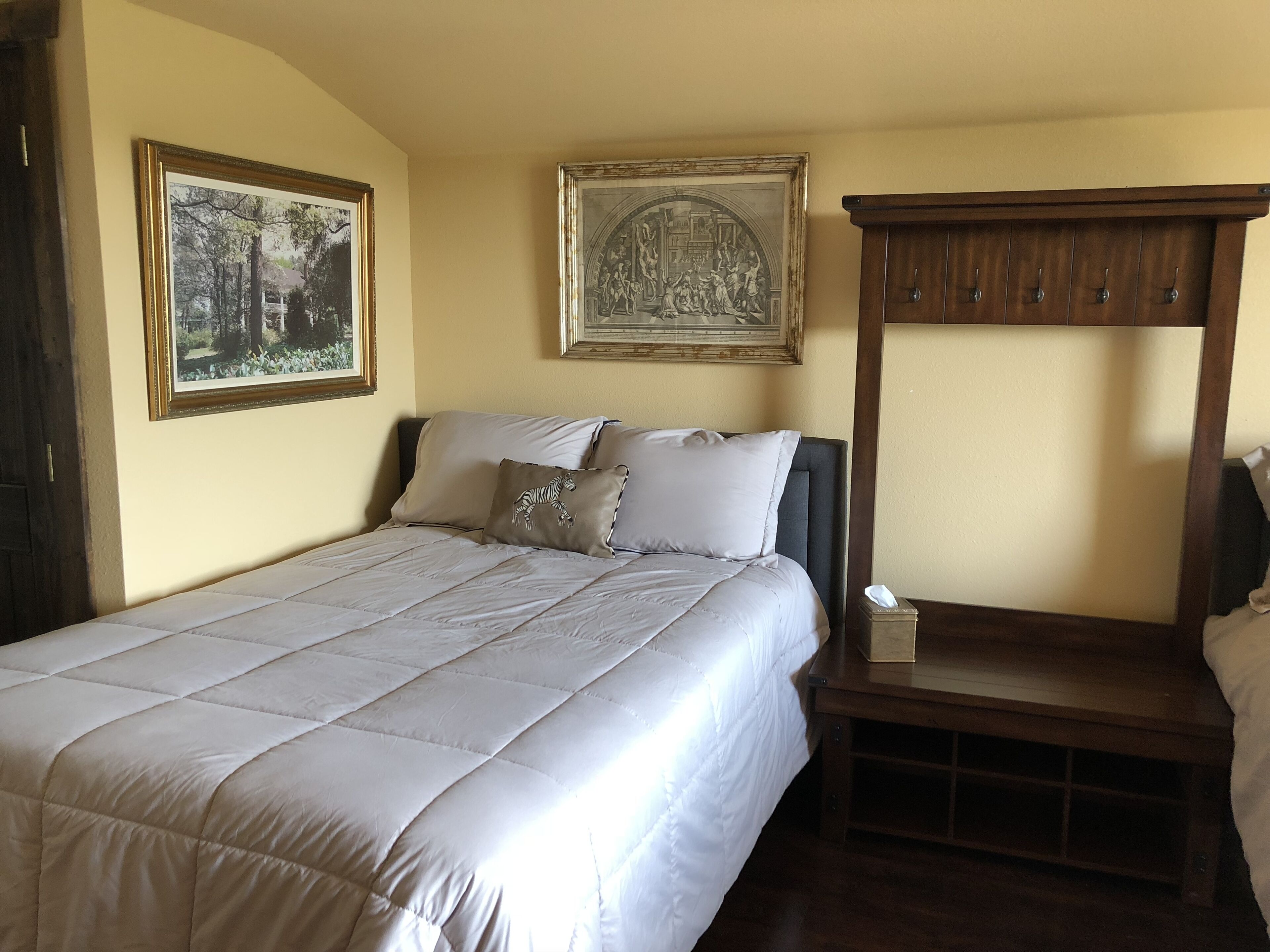 A neatly made bed with a quilted white duvet, two pillows, and a zebra-themed pillow. There is a wooden bedside table with a tissue box and a small lamp. Two framed artworks are hung on the wall above the bed.