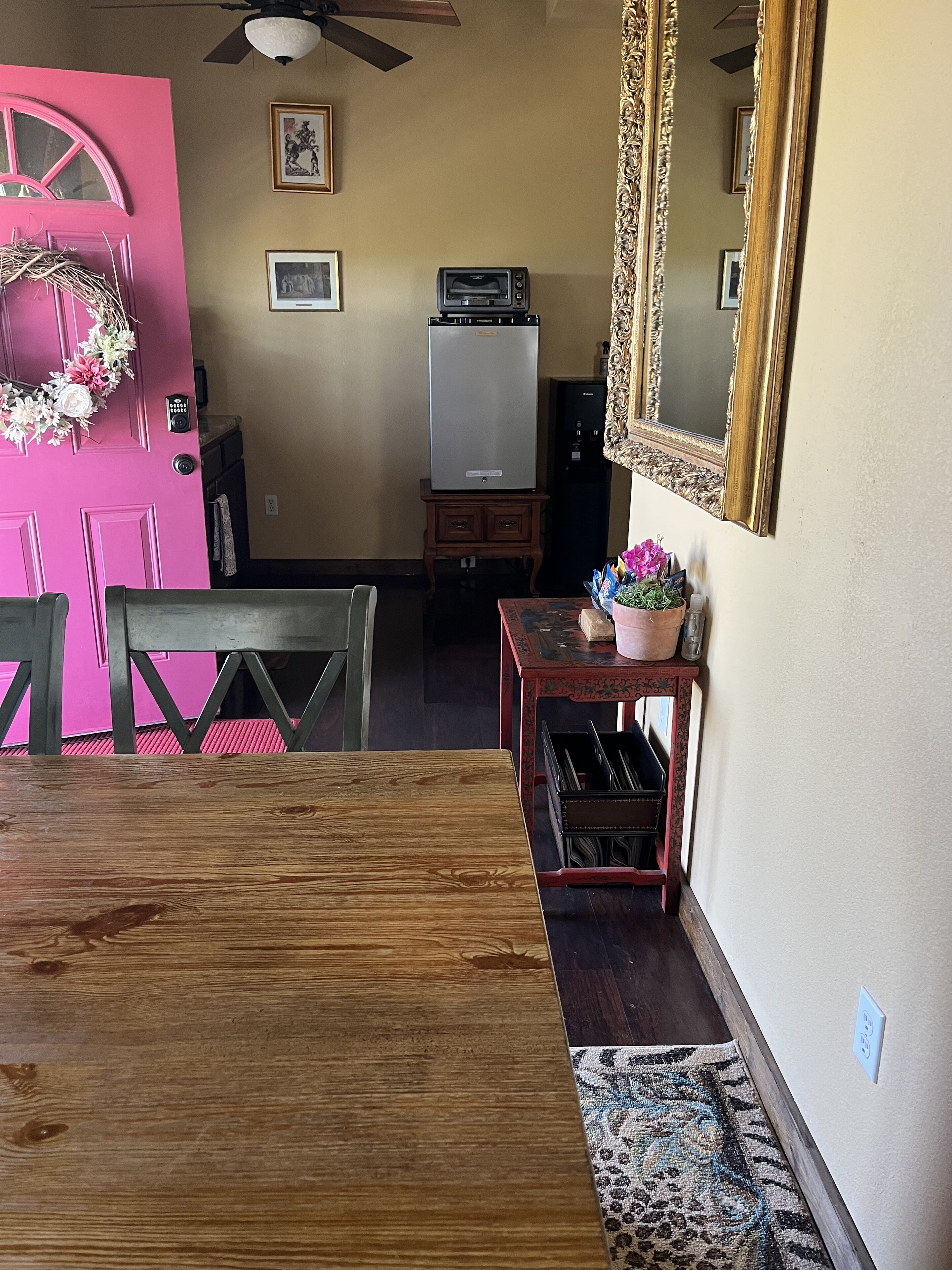 Un coin repas avec une table en bois, des chaises, une porte rose, un réfrigérateur, un petit meuble et un miroir au mur.