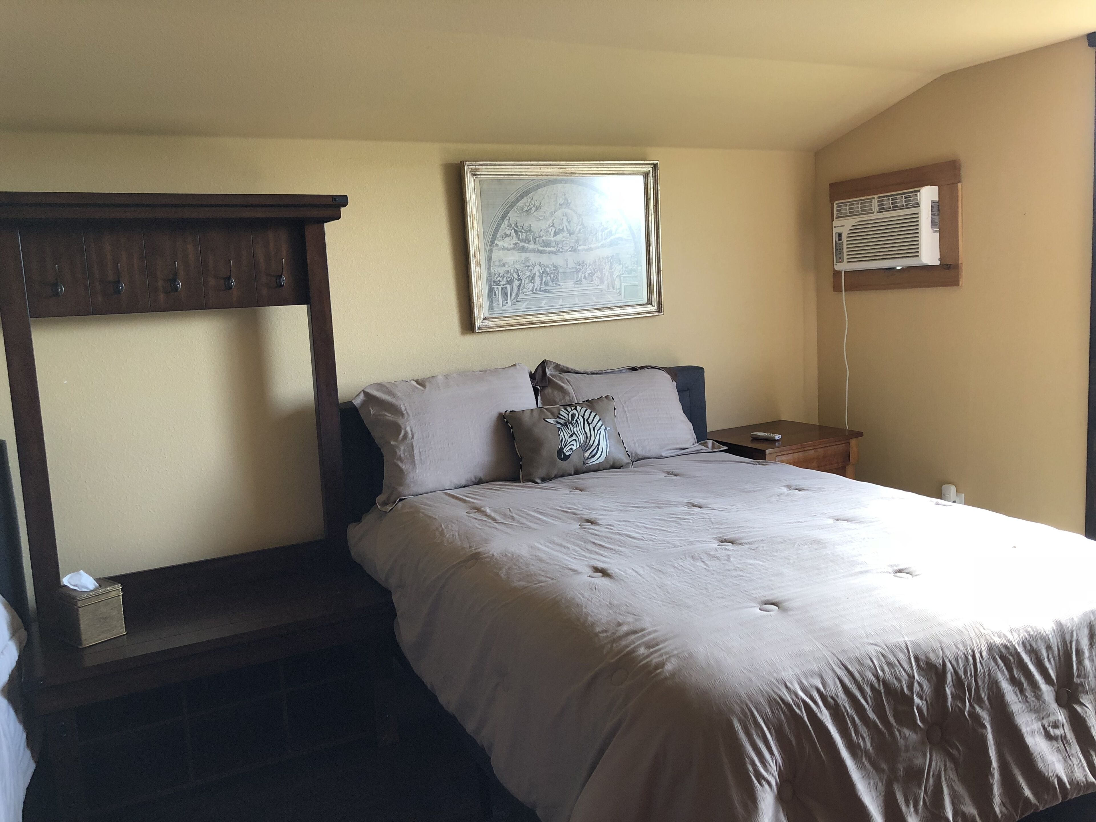 A neatly made bed with a headboard, a bedside table, a remote control, and a framed picture on the wall.