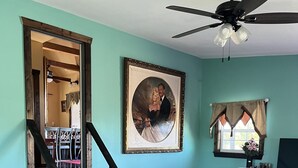 Una sala de estar con una escalera, un ventilador de techo, un retrato enmarco y una ventana con cortinas.