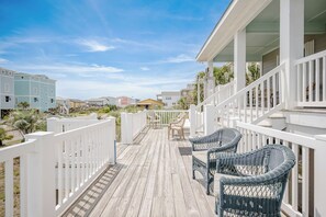 A wooden deck with wicker chairs, white railings, and a view of neighboring buildings. - Beach Joy: Bright, Ocean View Home Close to Beach, Splash Pad (Oak Island)