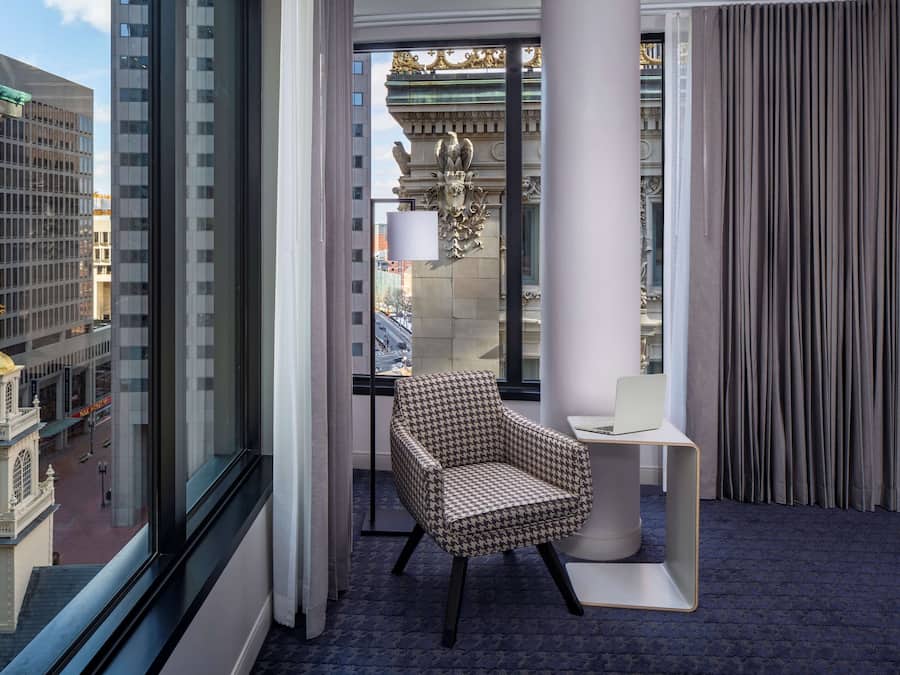 Habitación de esquina con un ventanal grande, un sillón estampado, una mesita blanca y vistas a un edificio histórico.