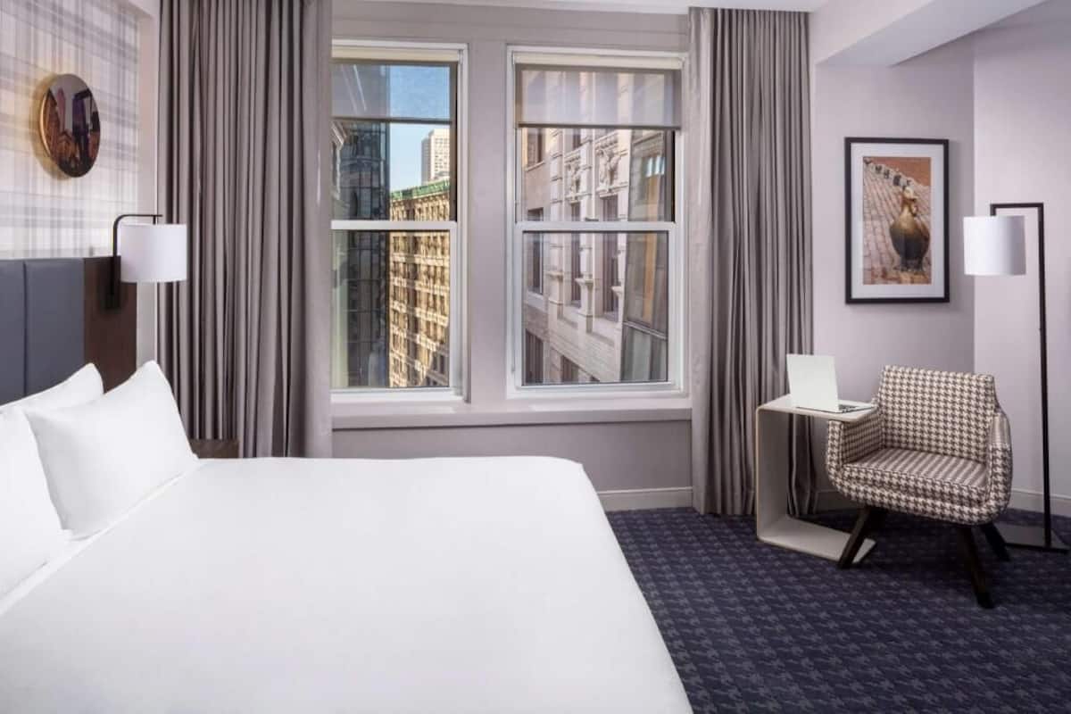 A hotel room with a large bed, two windows, a chair, and a lamp.