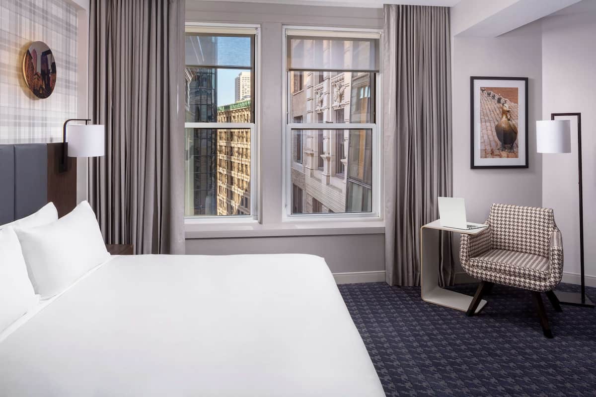 A hotel room with a large bed, two windows with curtains, a chair, a desk, and a framed picture on the wall.
