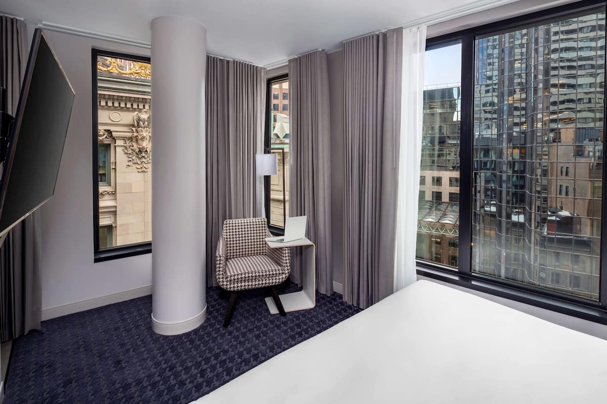 A hotel room with a bed, a desk with a laptop, a chair, a TV, and large windows with a city view.