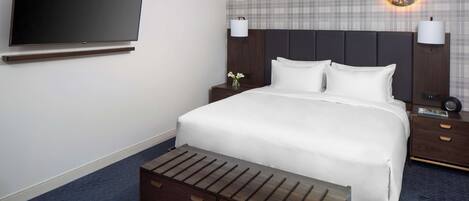 Una habitación de hotel con una cama grande, un televisor de pantalla plana montado en la pared, una mesita de noche con lámpara y una mesita auxiliar con reloj.