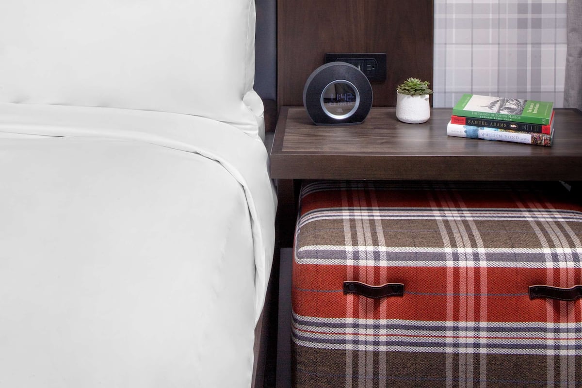 A neatly arranged hotel room with a bed, a bedside table with a lamp, a clock, and a small plant.