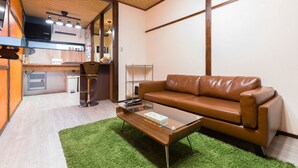 A compact living space with a brown leather sofa, a glass coffee table, and a small kitchen area with a microwave and a sink.