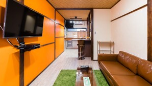A compact living space with a TV mounted on a wall, a sofa, a small kitchen area, and a dining table.