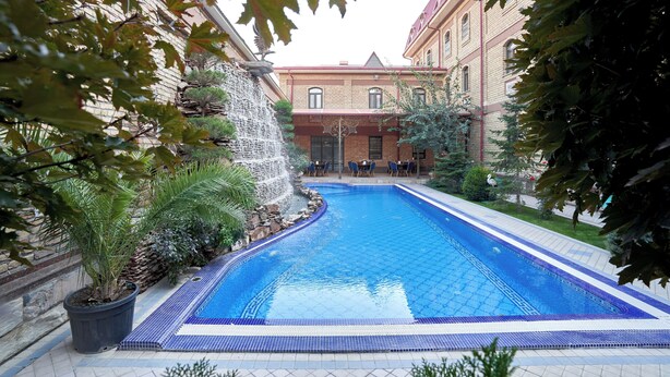 A swimming pool with a waterfall feature, surrounded by potted plants and a brick building in the background. - Rakat Plaza (Tashkent)