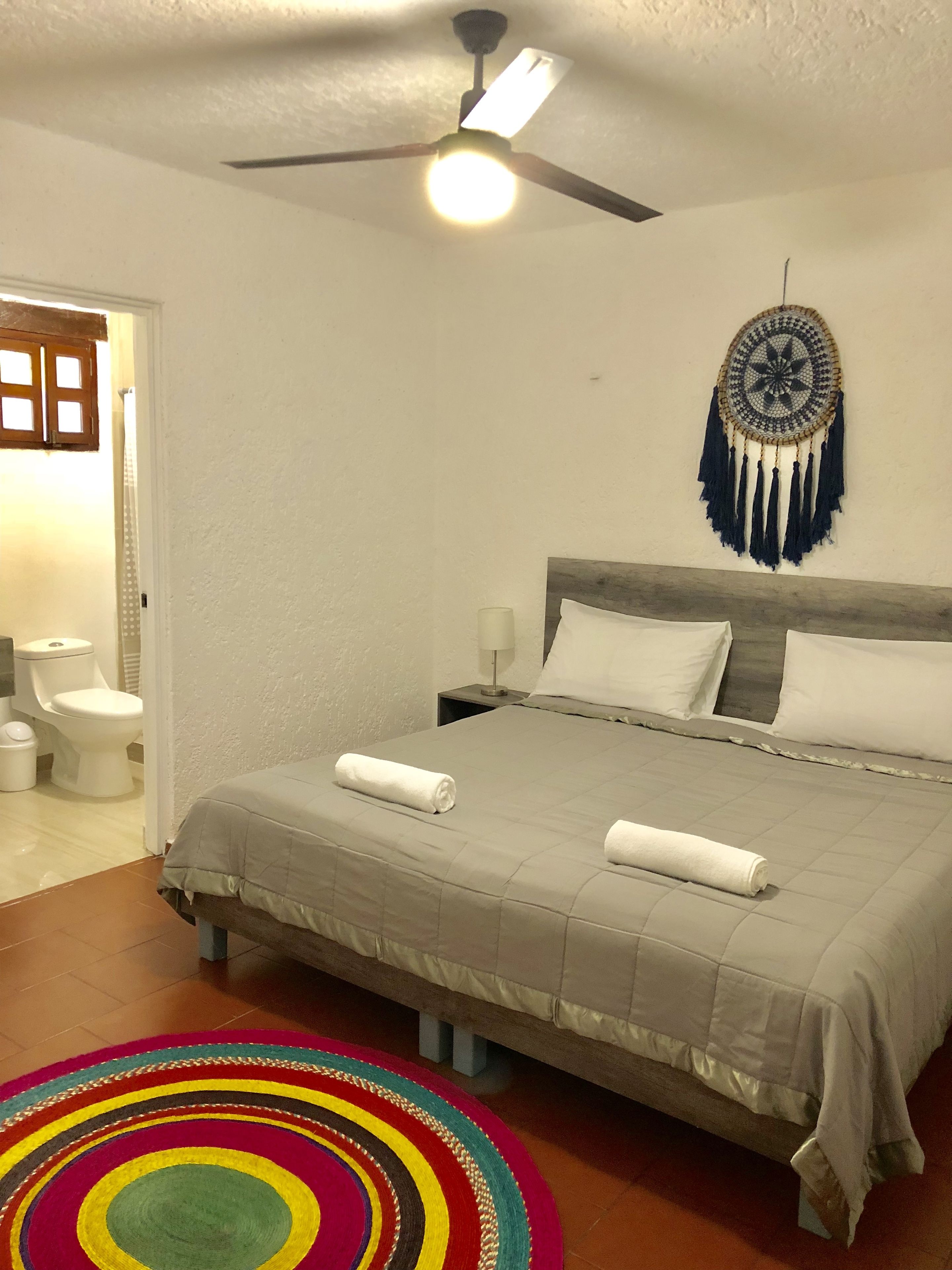 A bedroom with a bed, a ceiling fan, a nightstand, a wall-mounted dreamcatcher, and a colorful rug.