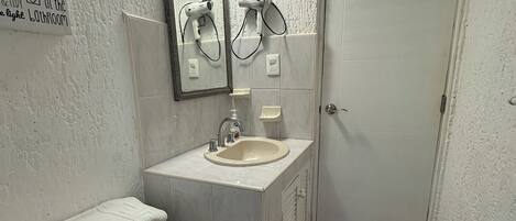 Un baño pequeño con inodoro, lavamanos y espejo. Hay un cartel en la pared y una puerta a la derecha.