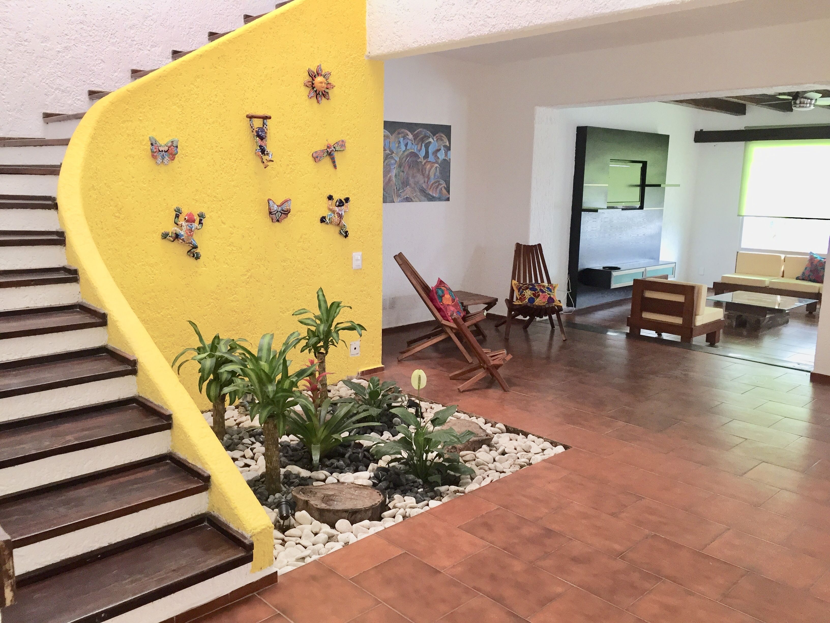 Un escalier avec un mur jaune, des décorations murales représentant des papillons, une chaise en bois, une table basse en verre et un coin salon avec un canapé et des coussins.