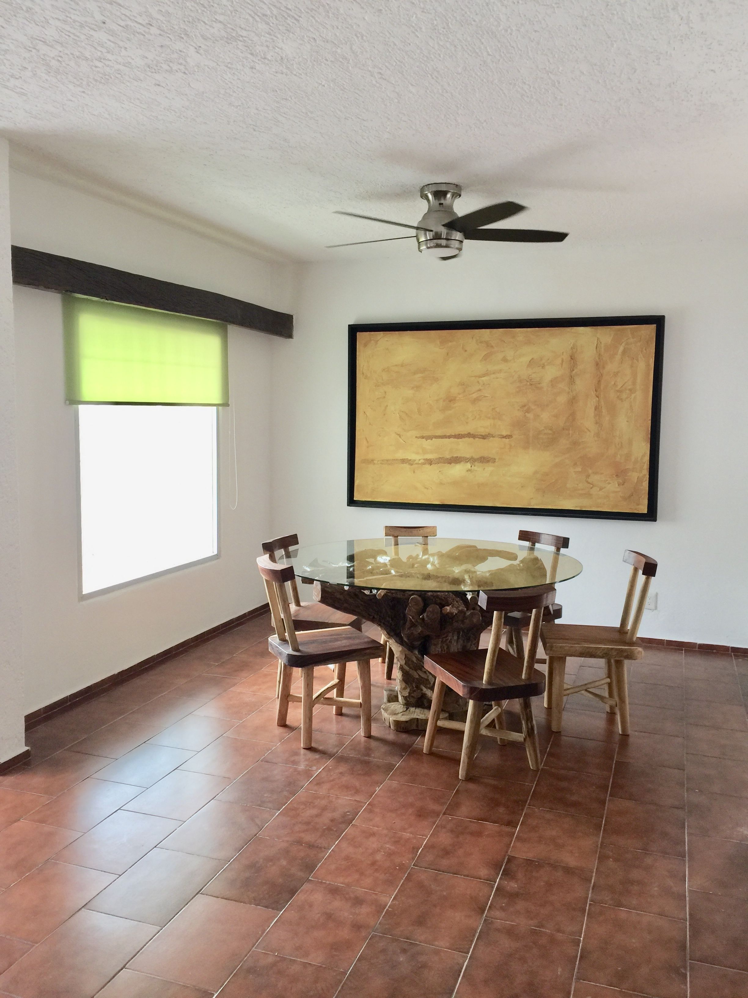 A dining area with a glass-top table surrounded by wooden chairs, a ceiling fan, a large abstract painting on the wall, and a window with a green blind.