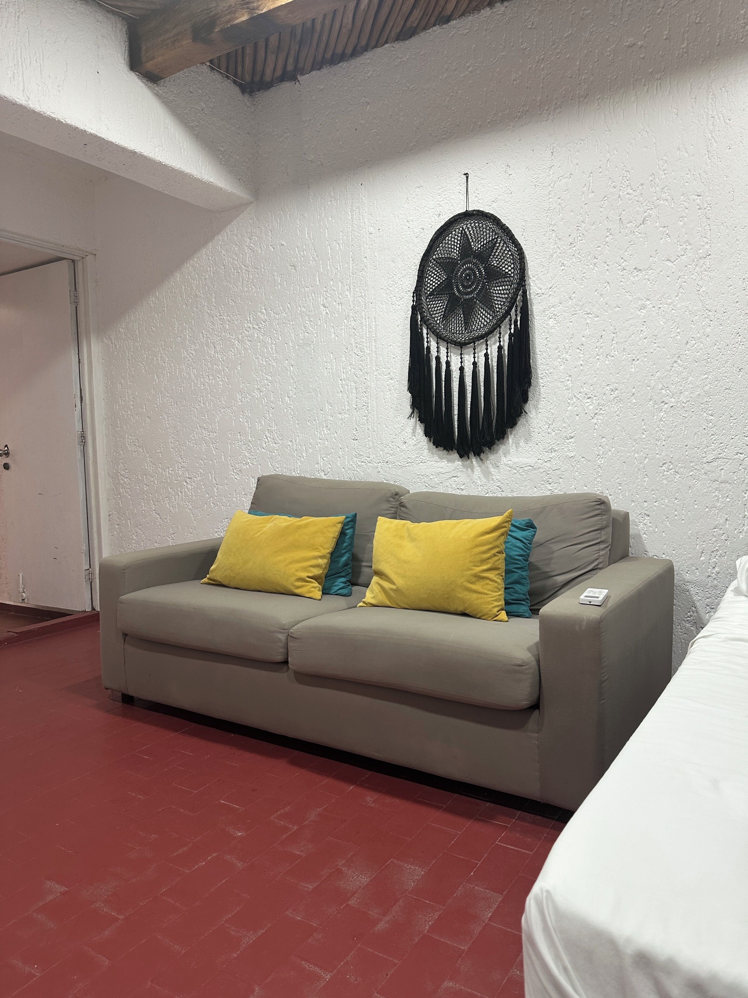 Ein Zimmer mit grauem Sofa, gelb-blaugrünen Kissen, einem schwarzen Traumfänger an der Wand und einem weißen Bett.