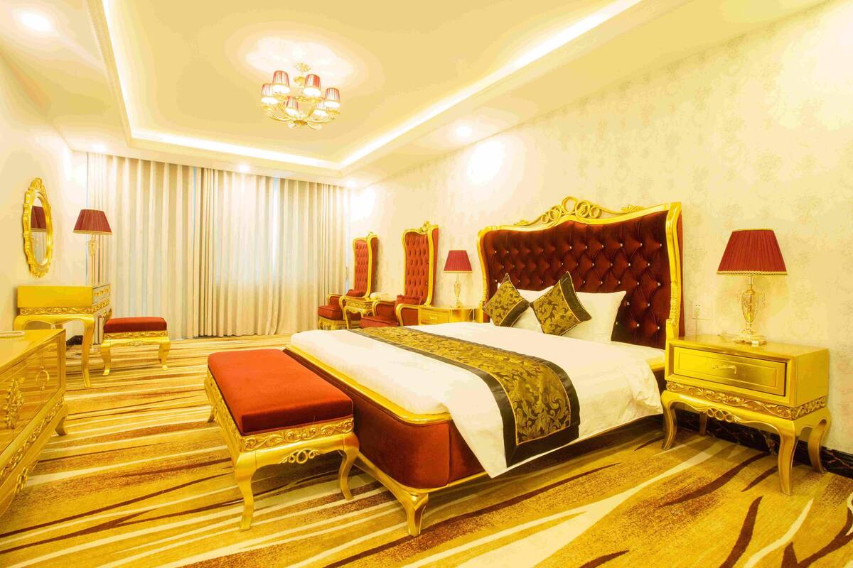 A luxurious hotel room with a large bed, ornate headboard, and a striped carpet.