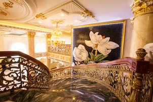 Uma escadaria ornamentada com piso de mármore e corrimãos dourados, que leva a um quarto com uma grande pintura floral e um candelabro.