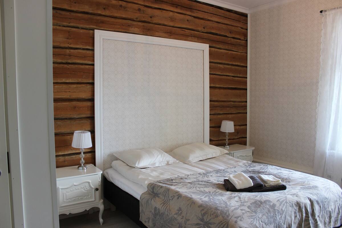 Een slaapkamer met een houten wand, een bed met bloemenbeddels, twee nachtkastjes met lampen en een bijzetmeubel.