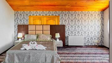 Habitación de hotel con techo de madera, papel tapiz estampado, cama con colcha, dos lámparas de noche y un radiador.