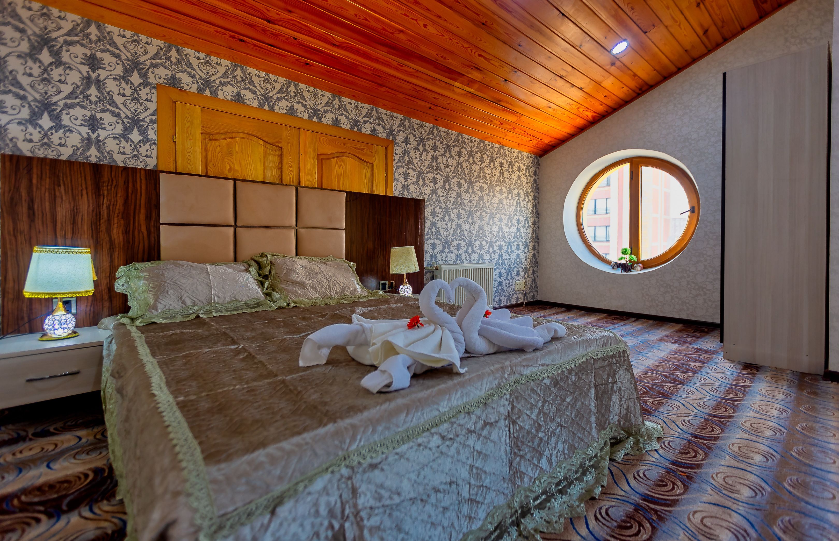 Un dormitorio con techo de madera, papel tapiz estampado, una cama grande con colcha marrón, dos lámparas de noche, un ventanal redondo y un radiador.