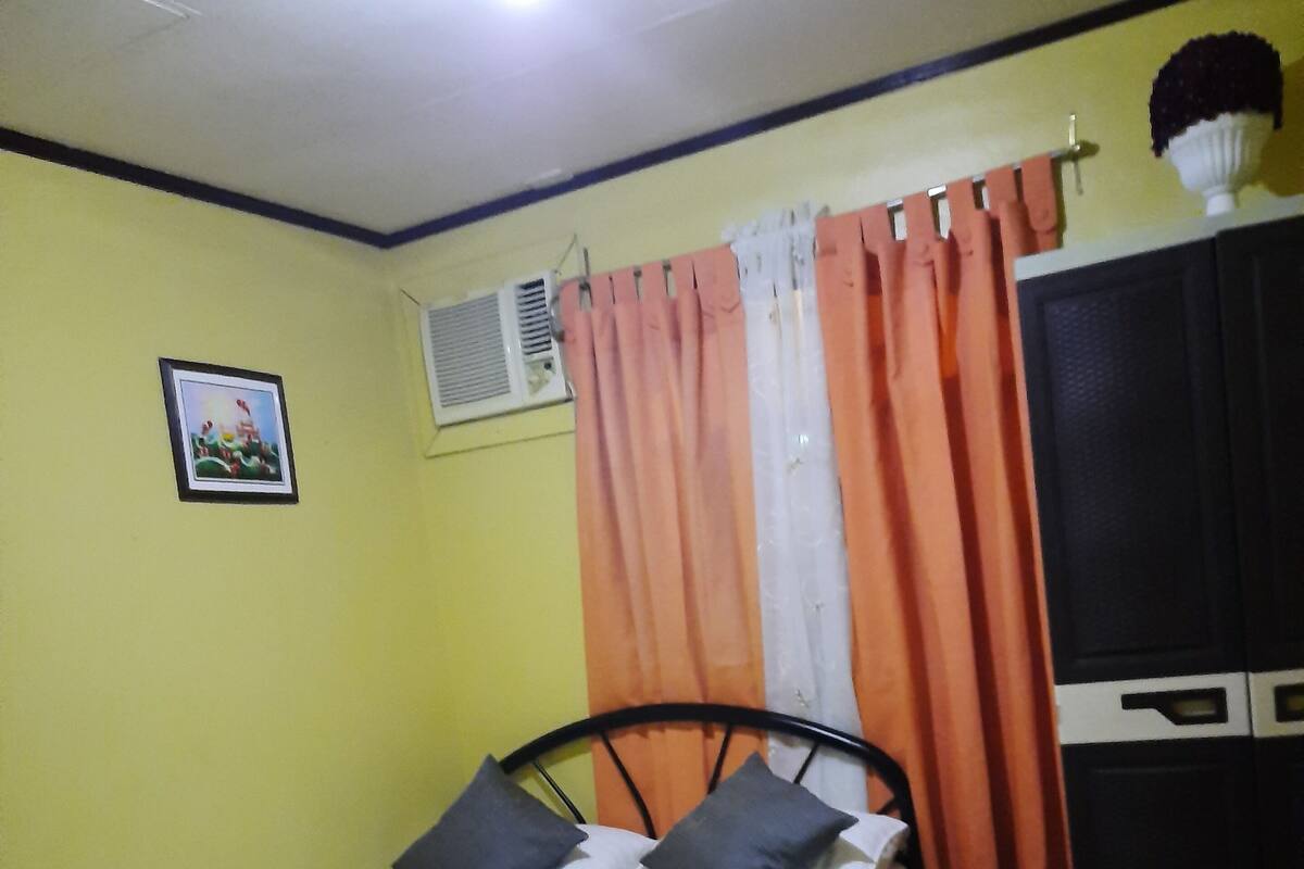 A single bed with a metal frame and two pillows, a small air conditioning unit, a television cabinet, and a patterned floor.