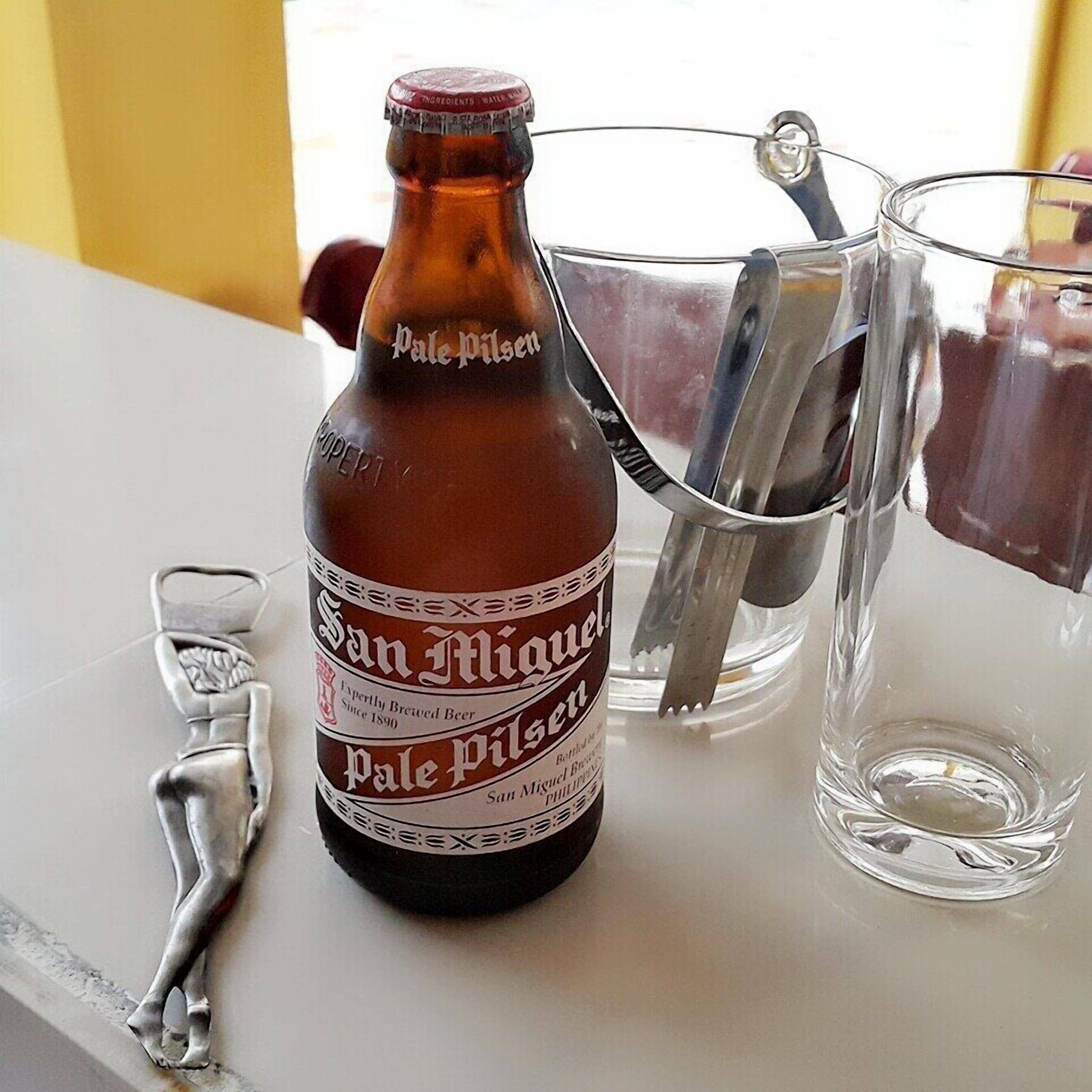 Una botella de cerveza San Miguel Pale Pilsen, un abrelatas de metal y dos vasos sobre una mesa.