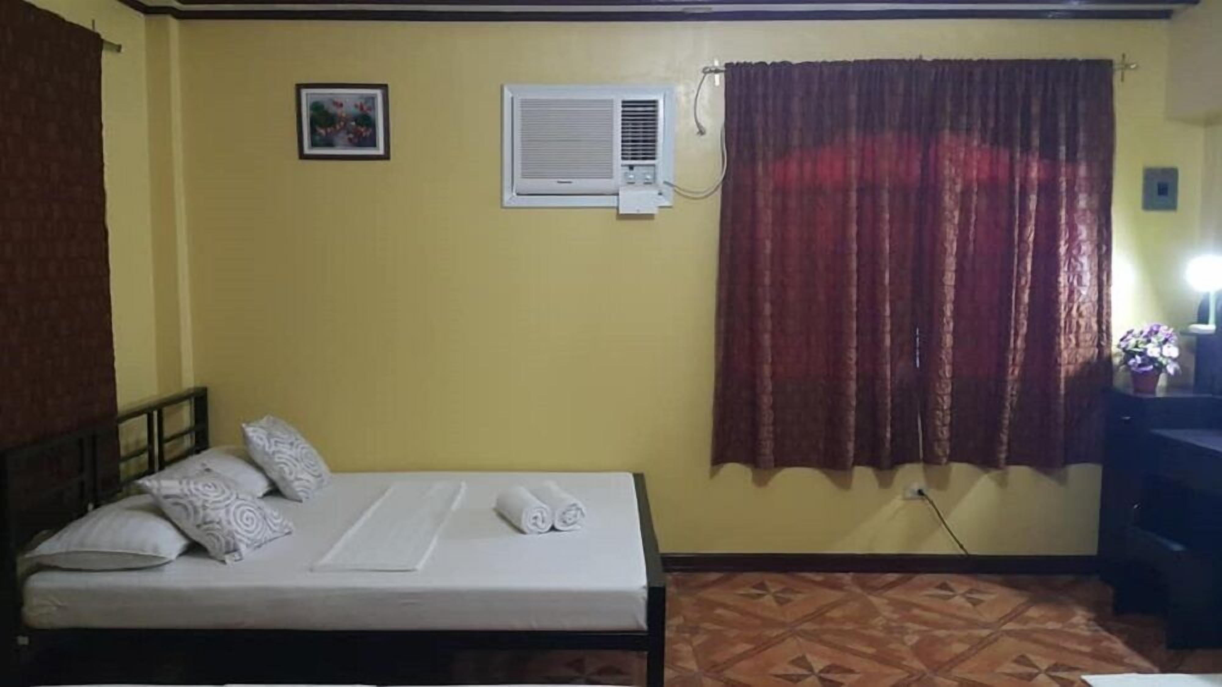 Een slaapkamer met een bed, een wandairconditioner, een raam met gordijnen en een klein tafeltje met een lamp.