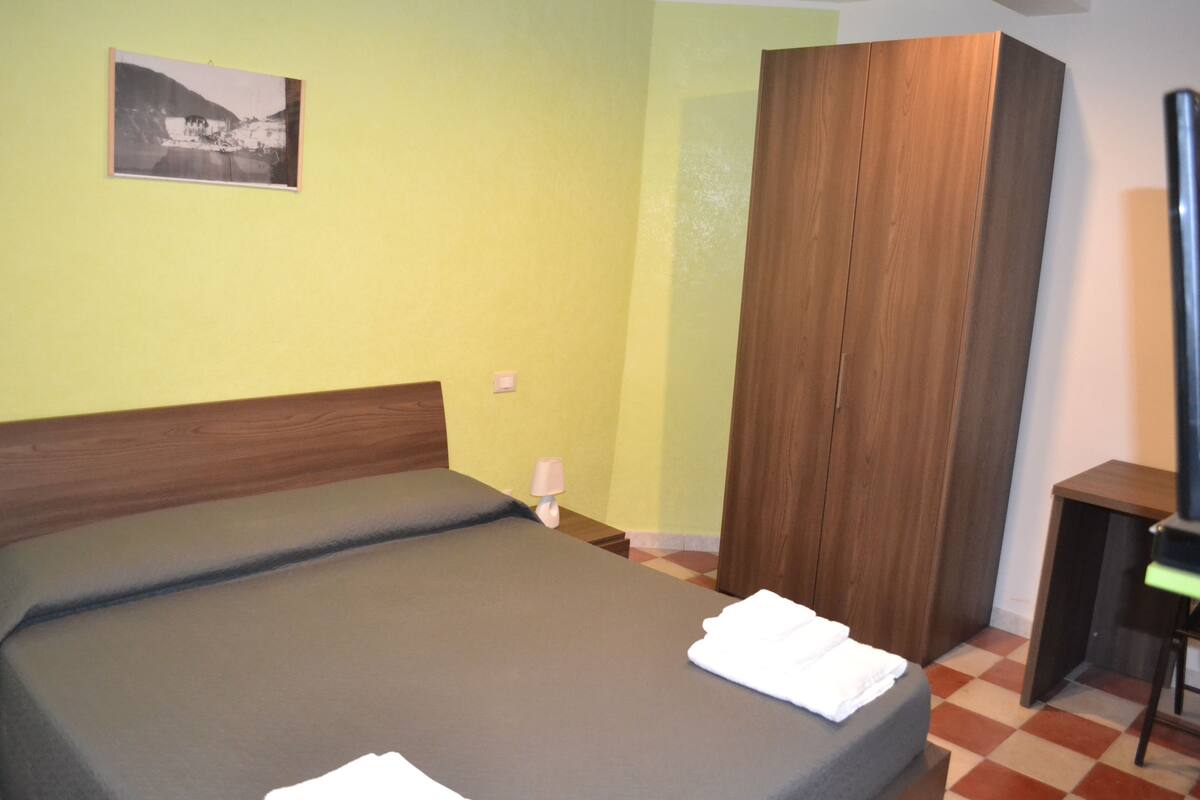 A room with a bed, a wooden wardrobe, a small table, and a wall-mounted lamp.