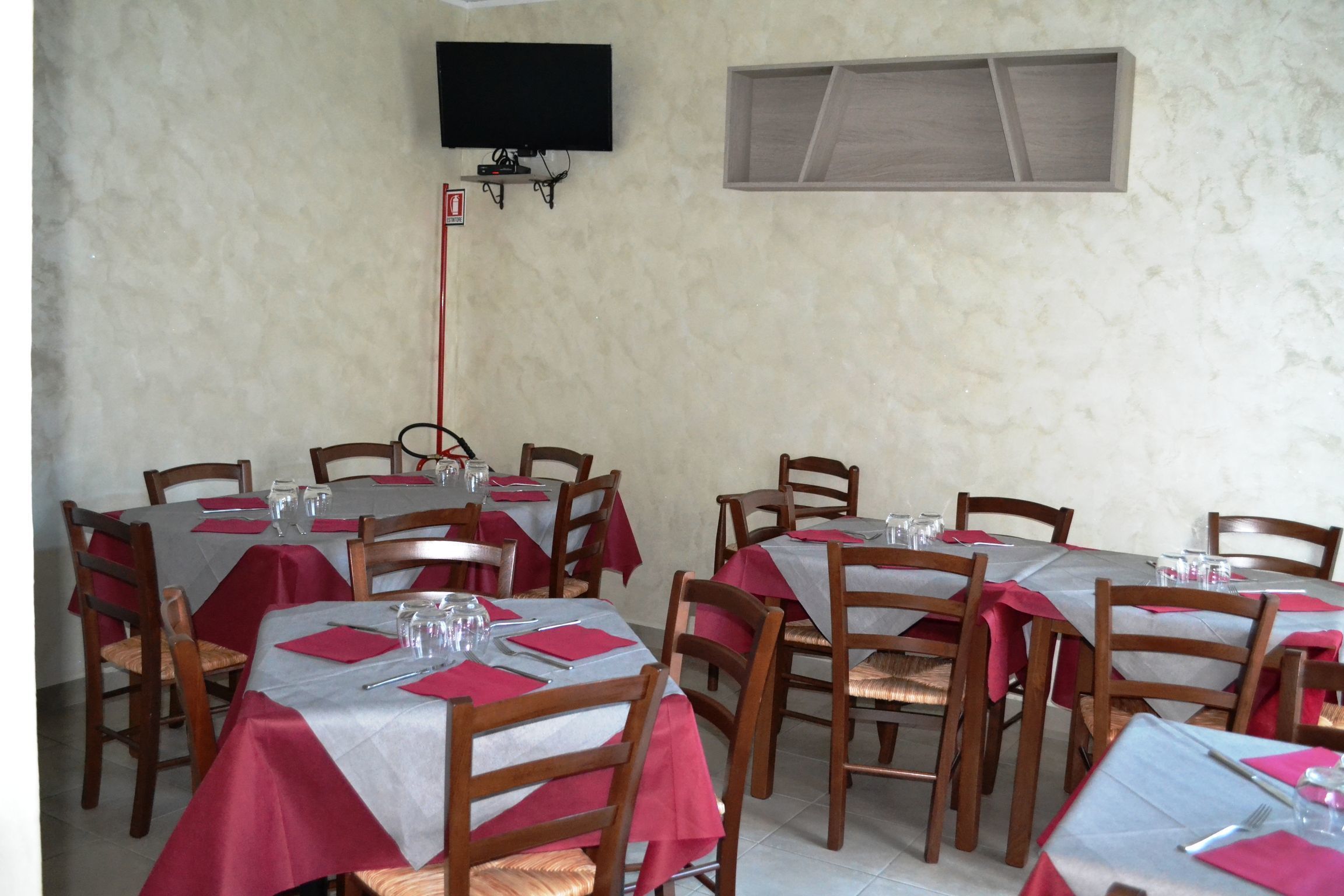 A restaurant with multiple tables covered with red and white tablecloths, wooden chairs, and a television mounted on the wall.