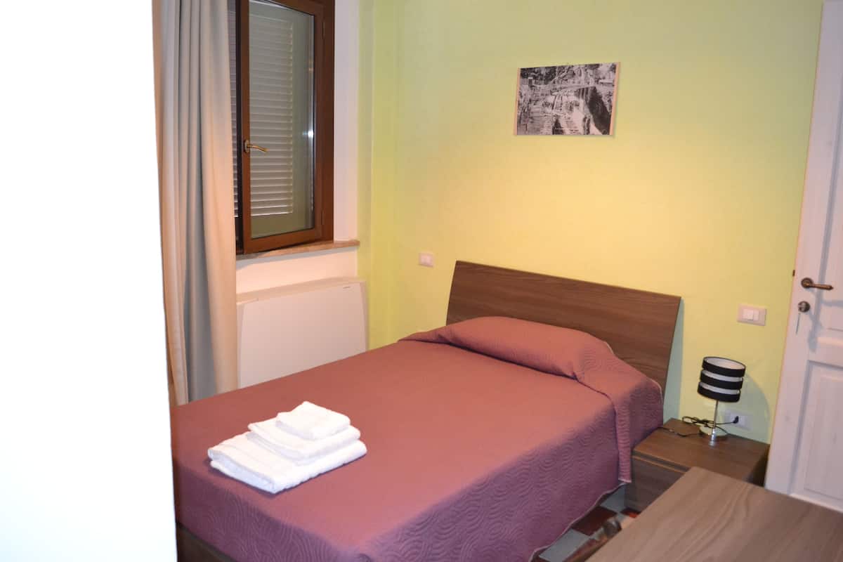 A neatly made bed with a pink bedspread and white towels, a wooden headboard, a small bedside table with a lamp, and a picture on the wall.