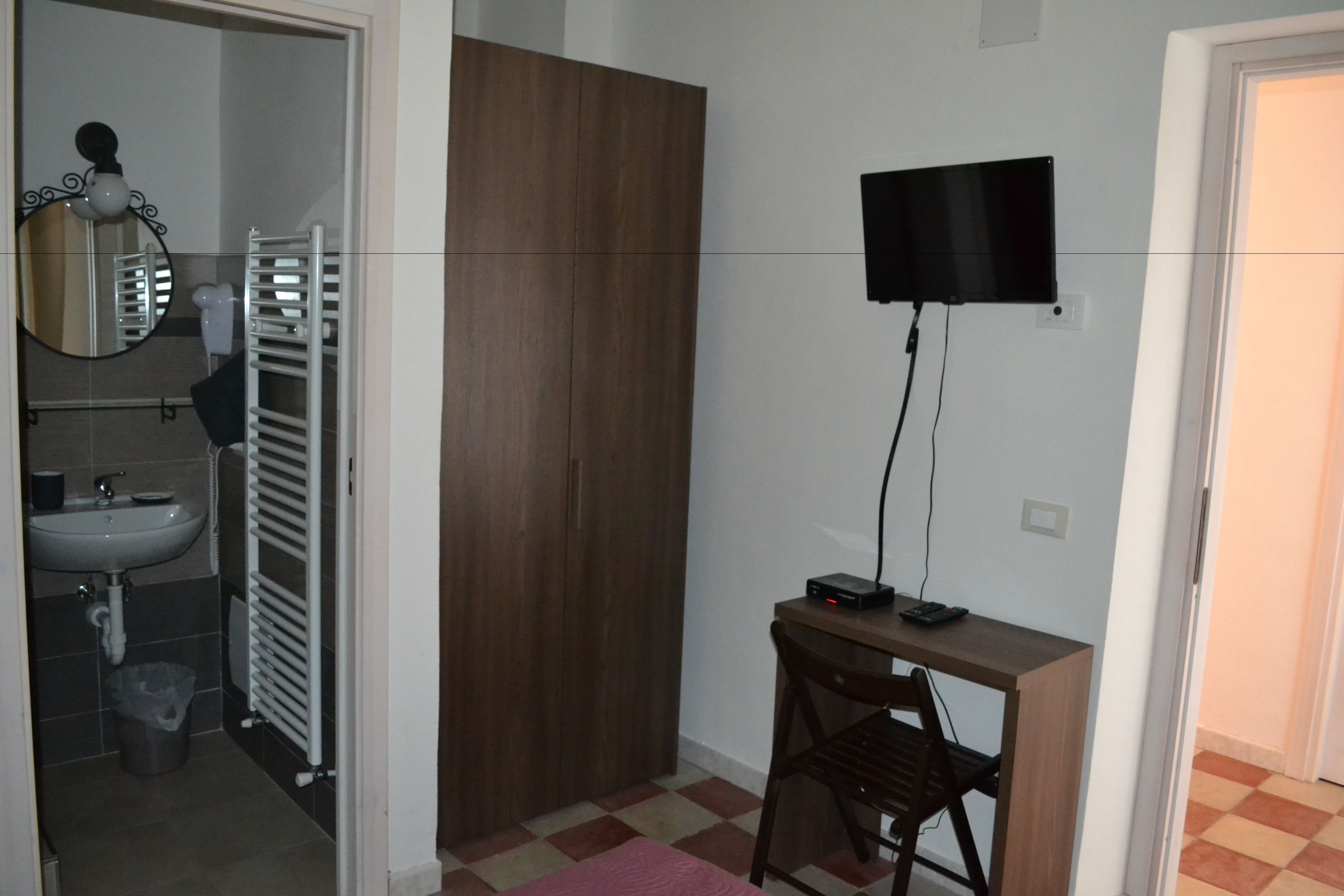 A small room with a wooden wardrobe, a desk with a computer, a mirror, and a sink.