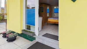 Un edificio amarillo con puerta de madera que da a una habitación con cama y una mesita. Hay plantas en macetas afuera.