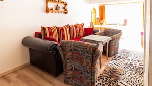 A living room with a sofa, two armchairs, a patterned rug, and a wall-mounted shelf with decorative items. - Holiday apartment - FH VIL 0062 (Patzig)